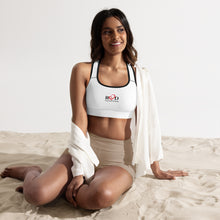 Load image into Gallery viewer, image of a slim brown model sitting on sand wearing cropped white padded sports bra with racer back, which has contrast dark stitching and black trim around the neck and arm holes, together with a Red Valenteen logo center front. Teamed with a draped over shirt, and beige lycra shorts.