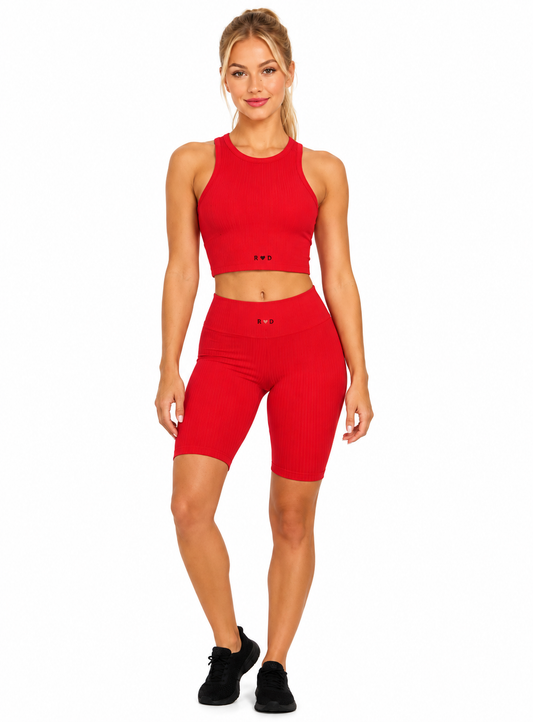 A fitness model wearing the two red pieces from the Red Valenteen collection . The red racerback tank top with the small Red Valenteen logo centered on the waistline, and the red mid length shorts and black sneakers. On a white background.