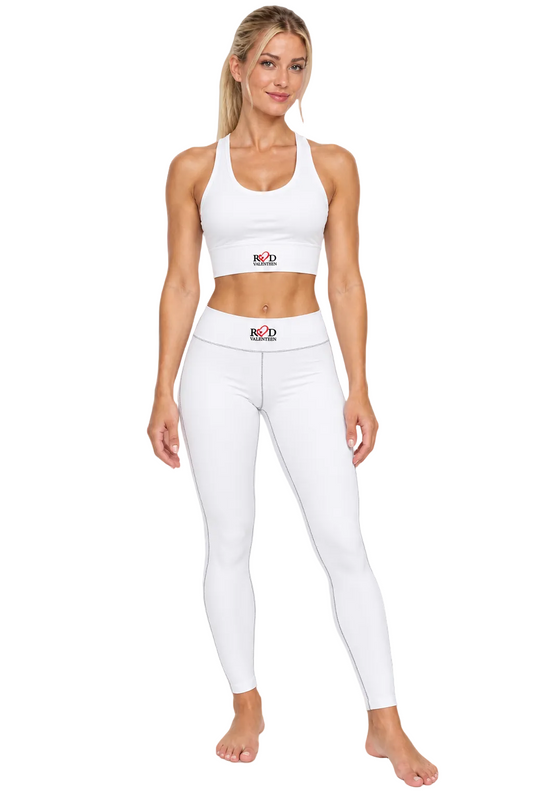 A woman modeling a white athletic two-piece set consisting of a sports cropped racerback top and high-waisted leggings, both featuring a small red and black logo, Red Valenteen on the front. The background is a clean, solid white.