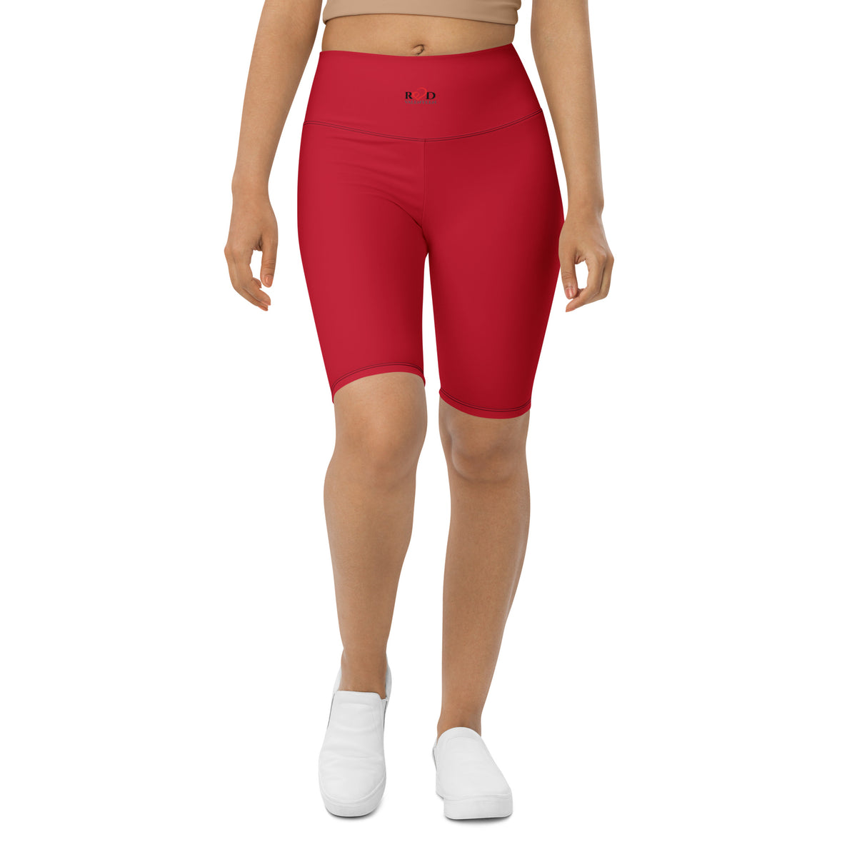 A model wearing bright red high-waisted biker shorts, mid length above knee, paired with a beige crop top and white slip-on shoes, shown against a plain white background.
