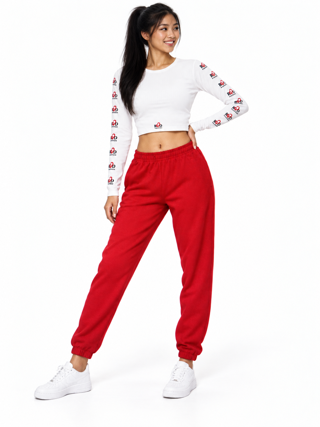 A woman modeling a two-piece outfit consisting of a white long-sleeve crop top with a 'Red Valenteen' pattern down the sleeves, paired with bright red jogger sweatpants and white sneakers, against a plain white background.