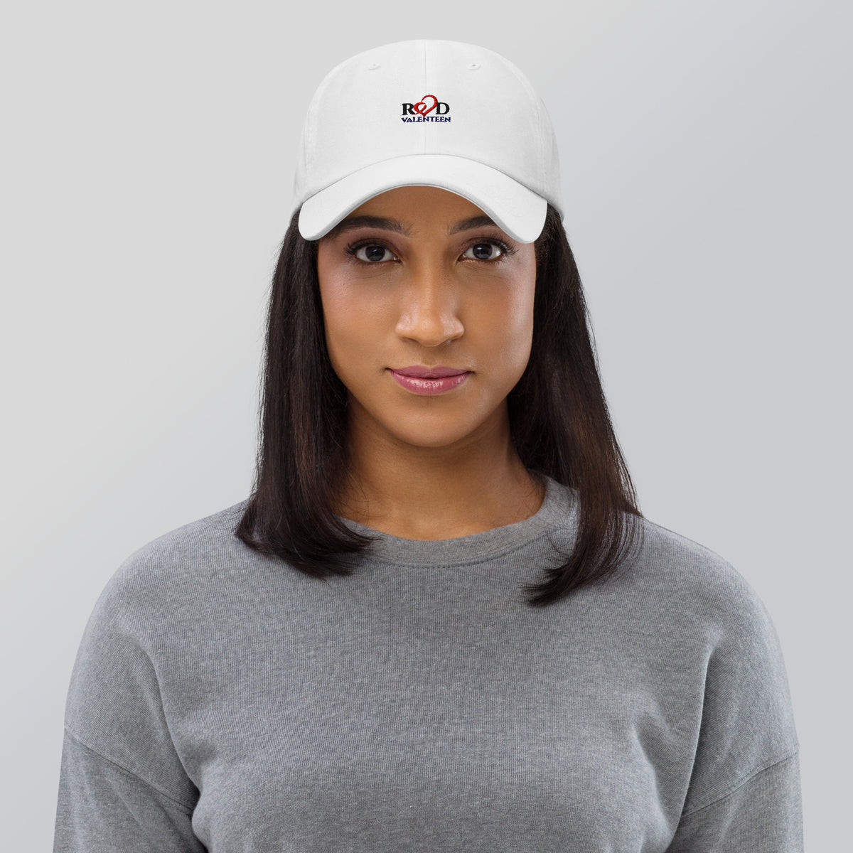 Women wearing white Red Valenteen logo cap facing the camera with a grey tshirt, shoulder length black hair, with a white background.